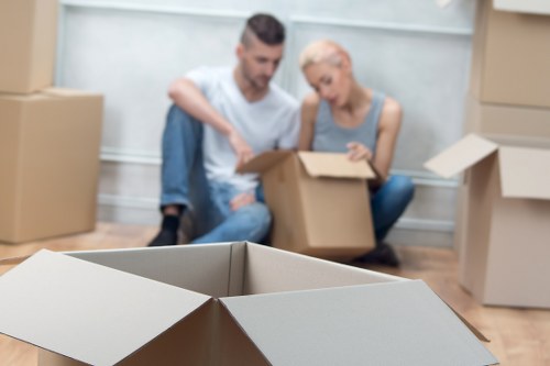 Two individuals, a man with dark hair and a woman with short blonde hair, sit on a hardwood floor inside a residential property surrounded by several open and closed cardboard moving boxes. The man is wearing a light blue T-shirt and jeans, and the woman is dressed in a sleeveless light grey top. They are engaged in packing or unpacking, with one of them holding a cardboard box lid. The boxes, made of brown corrugated cardboard, vary in size and are positioned in the foreground and background, some stacked or partially collapsed. The environment appears to be a clean, well-lit interior, possibly a living room or hallway, with a white wall behind them. The scene suggests a house relocation process typical of house removals services, with no visible furniture, appliances, or other household items, only the packing materials. The focus is on the collaborative effort of the moving process, capturing a moment within a house in Worcester Park as part of professional removals.