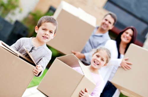 A family of four, including a young boy and girl, along with a man and woman, are outside a residential house during daylight hours, engaged in a home moving process. The boy in the foreground is holding a large cardboard box, which appears to be filled with household items; the box is plain brown with visible flaps. The girl next to him is smiling and also holding a similar cardboard box. The man, standing behind them, is carrying a medium-sized cardboard box, and the woman next to him is supporting a larger box, slightly behind the others. All boxes are made of brown corrugated cardboard, showing signs of handling, and contain various household belongings. The background includes a modern house with large windows and a garden with neatly trimmed grass, suggesting a suburban area, possibly in London or nearby. The scene is well-lit with natural daylight, emphasizing the brightness of the outdoor environment, and reflects a typical house removal activity coordinated by Watson Removals London, highlighting their service in the local area around postcode SE or SW London.