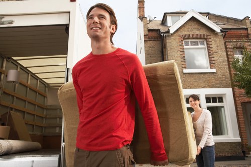 A young man in a red long-sleeve shirt is outdoors in a residential street in Hampton Hill, carrying a beige upholstered armchair with a wooden frame over his shoulder, part of a house removal process. Behind him, a woman in a striped top stands near a brick house with a white-framed window, observing as they prepare to load furniture into a removals van parked in the driveway, which is partially visible with an open tailgate and packing materials inside. The scene includes typical residential house features such as brickwork, a small front garden, and a tiled roof, with natural daylight illuminating the area, suggesting a clear day. This setup indicates a professional move by Watson Removals London in the Hampton Hill area, with household furniture and items being carefully handled and loaded for transport.