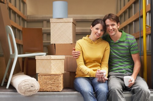 A young couple sits closely together on the floor of a residential garage or storage area, surrounded by packing boxes and household items. The woman wears a yellow hoodie and jeans, and holds a mug, smiling at the camera. The man, dressed in a green and purple striped polo shirt and grey trousers, also smiles and holds a mug. Behind them, there are stacked cardboard boxes, some wrapped in packing paper, and a small blue roll of packing tape on top of a beige box. To the left, a white plastic chair leans against the wooden wall, and a rolled-up white carpet or rug is placed on the floor nearby. The environment appears to be a typical internal space used for storage during a house move, with natural lighting and an organised arrangement of household contents, reflecting the context of house removals in London or nearby areas.
