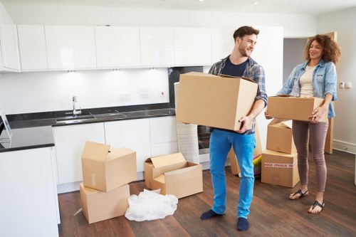 A young man and a woman are inside a modern kitchen, preparing to move belongings. The man, wearing a plaid shirt and blue jeans, is holding a large cardboard box close to his chest, smiling as he looks at the woman. The woman, with curly red hair, dressed in a light blue denim jacket and burgundy trousers, is carrying a smaller cardboard box. Several unpacked cardboard boxes and some crumpled packing paper are scattered on the dark wooden floor. The kitchen features white cabinetry, a black countertop, and a stainless steel sink. Natural light illuminates the scene, with a spacious, tidy interior typical of a residential home in London. This image exemplifies home moving activities undertaken by a professional removals service, such as Watson Removals London, highlighting careful handling of household items during a residential relocation within the area.