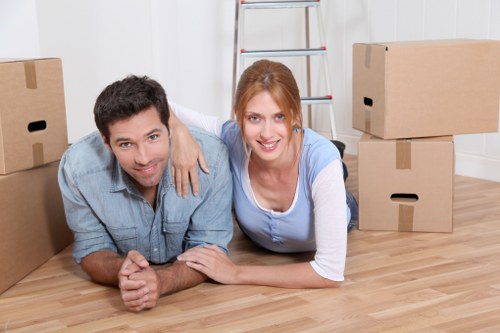 A man and a woman are lying on the wooden floor of a residential interior, surrounded by packed cardboard boxes of various sizes, indicating a house move. The man has dark hair and is wearing a light blue shirt, while the woman has red hair and is dressed in a light blue and white top. They are smiling at the camera, with the woman's arm draped over the man's shoulder. Behind them, a silver stepladder leans against a white-painted wall, and the room appears well-lit with natural or overhead lighting. The scene captures a moment during a professional house removal process, likely in the Motspur Park area, as part of Watson Removals Londonâ€™s local relocation services. The boxes, along with the plain white wall and wooden flooring, suggest it is a typical residential space ready for packing or unpacking, emphasizing the professional and organized approach of a removals company operating in the London area.
