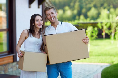 A smiling young couple standing outside a residential property in Soho, each holding medium-sized cardboard moving boxes. The woman has long dark hair and is wearing a white sleeveless dress, while the man has short light hair and is dressed in a white shirt and blue trousers. The background features a house with large windows, part of a garden with green grass and trees, and a clear, bright sky. They appear to be in the process of moving home, with the boxes possibly filled with personal belongings. The scene is well-lit with natural light, emphasizing a professional but friendly atmosphere typical of house removals in the local area, and subtly referencing the services of Watson Removals London in facilitating residential moves in Soho.