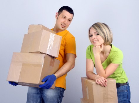 A male mover wearing a yellow t-shirt and blue gloves is holding two cardboard boxes stacked vertically, one on top of the other, in a well-lit indoor setting, likely a residential property in Leaves Green. Behind him, a female mover, dressed in a bright green top, is leaning on a table or surface, supporting a medium-sized cardboard box with her right arm, while her left hand rests on top of the box. The scene suggests a house removal process, with the movers prepared to transport personal belongings during a relocation. The background features a clean, neutral grey wall, and the lighting highlights the items and individuals clearly. The boxes are standard brown cardboard, with some taped edges visible, indicating packed household items ready for moving. This scene exemplifies professional house removals in the Leaves Green area, with focus on careful handling of furniture and boxes, characteristic of Watson Removals Londonâ€™s services for local moves within the area or nearby neighborhoods.