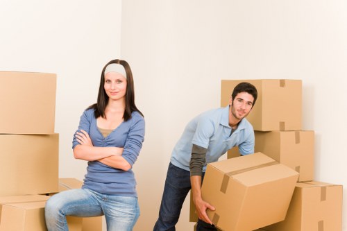 A young woman with long dark hair, wearing a blue long-sleeve top and light blue jeans, sitting on a small stack of cardboard boxes with her arms crossed and a neutral expression, in a plain, well-lit indoor space. Beside her, a young man with short dark hair, wearing a light blue shirt over a darker long-sleeve shirt and dark jeans, is kneeling slightly while holding a large cardboard box, preparing to place it on the ground. Surrounding them are several other cardboard boxes of various sizes, some stacked against a white wall, indicating a household or moving environment in Bellingham. The scene is clean, minimal, and focused solely on the relocation activity, underscoring professional house removals services typical of Watson Removals London, with a neutral background free of furniture or other household items, and the lighting suggests daytime interior conditions.