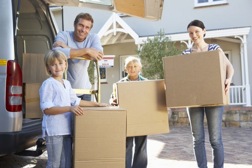 A family of four, including a man, a woman, and two children, are participating in a house removal on a residential street in Soho. They are standing outside a grey van with an open rear door, which is filled with cardboard boxes and household items. The man is smiling and leaning on the van, while the woman holds a large cardboard box. The girl, with blonde hair, is holding a smaller box and smiling at the camera, and the boy, also blonde, stands between the boxes, looking forward. The street is lined with houses featuring white porches and gardens, with a clear sky above. The family appears to be engaged in packing or unloading, capturing a routine moment during a professional removals service provided by Watson Removals London. The scene focuses on the early stages of relocating within Soho, with the family's cheerful expressions and the lifting of moving boxes reflecting a positive transition during a home change process, supported by local house removal experts.