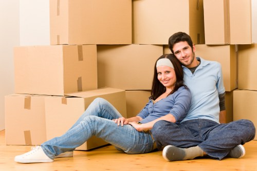 A young couple sitting on a wooden floor surrounded by large cardboard boxes and packing materials, with some boxes stacked against a wall in the background, suggesting they are in a residential interior preparing for a house move. The woman has long dark hair and is dressed in a blue top and jeans, while the man has short dark hair and is wearing a light blue shirt and dark trousers. The scene is well-lit with natural light, conveying a sense of preparation and organization typical of a house removal process in Mill Hill, as handled by Watson Removals London. The boxes appear sealed and ready for transport, with the couple seated comfortably, smiling, indicating they are in the midst of their moving arrangements within their home environment.