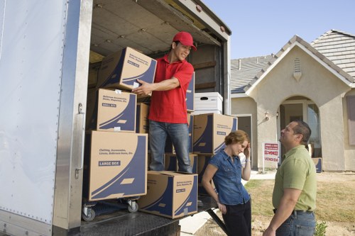 A moving crew member wearing a red cap and red T-shirt is inside a moving van, stacking and arranging large cardboard boxes with blue and white labels. The boxes are placed on a dolly and on the van's floor, and some are stacked vertically. Outside, a woman with red hair tied back, wearing a dark blue shirt, stands on the ground near the van, talking to a man dressed in a light green polo shirt and glasses, who is listening attentively. They are standing on a residential street in Cranham, with a light-colored house featuring a tiled roof and a small front garden visible in the background. The scene appears to be during daytime with clear weather, providing natural lighting that highlights the textures of the boxes, the clothing, and the surrounding environment. This image illustrates a typical house removal process in a suburb of London, with the Watson Removals London team engaged in loading household items for a local move.