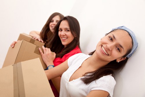 Three young women with light skin and dark brown hair are standing close together indoors in a bright, neutral-colored room, smiling at the camera. The woman in the foreground, wearing a white top and a blue headband, is holding two cardboard boxes, one with visible packing tape, suggesting they are in the process of a house removal or move. Behind her, a woman in a red top is also holding a box, while the third woman, partially visible, appears to be assisting or preparing to carry items. The scene reflects a scene of house removals with a focus on packing and moving household belongings, such as cardboard boxes and casual clothing, in a residential environment. The minimal background and soft lighting emphasize the collaborative effort involved in moving house services in Hampstead Garden Suburb, as provided by Watson Removals London.