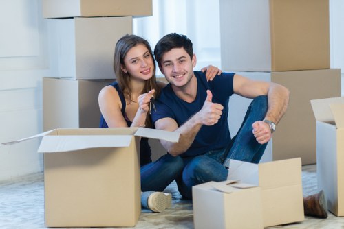A young couple sits on the floor inside a residential property surrounded by open cardboard boxes and packing materials, indicating they are in the process of packing or unpacking. The woman has long brown hair, is wearing a navy sleeveless top, and is smiling while leaning close to the man, who has short dark hair, is dressed casually in a dark blue T-shirt and jeans, and is giving a thumbs-up gesture. Behind them, large, stacked cardboard boxes are visible against a light-colored wall, with some boxes open showing their contents, and others sealed or awaiting packing. The natural light coming through the window illuminates the scene, highlighting the texture of the cardboard and the casual atmosphere of a house removal or relocation in the Creekmouth area. This scene reflects a typical household move, with a focus on the process of packing or unpacking in a residential setting, as might be supported by Watson Removals Londonâ€™s services in the local area.