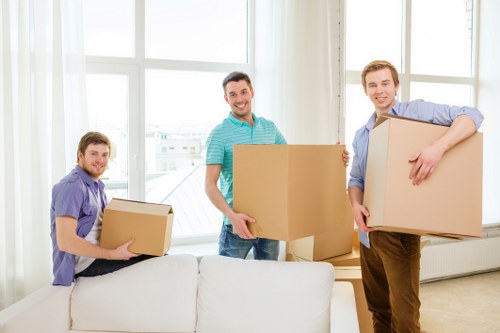 Three men with light skin are indoors in a bright, modern living room near a large window with white framing, during a house move in London. They are smiling and holding cardboard boxes of varying sizes, which are made of brown kraft material with visible flaps and tape. The man on the left, wearing a purple shirt, is seated on a white sofa with a soft fabric texture, holding a medium-sized box close to his chest. The man in the middle, dressed in a turquoise and white striped T-shirt, is standing, carrying a large box with both hands, positioned centrally in the room. The man on the right, in a light blue shirt and brown trousers, is standing behind the sofa, holding an even larger box with one arm, which partially obscures his outfit. The background reveals a tidy, well-lit space with cream walls, and the scene suggests an organized and careful packing process typical of professional removals in the London area, with a focus on efficient moving services by Watson Removals London. The environment appears clean and residential, emphasizing a professional, yet casual atmosphere conducive to house relocations.