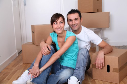 A smiling young couple sitting on a wooden floor inside a residential property in Archway, with several large cardboard moving boxes stacked behind them. The woman, wearing a bright blue top and jeans, is seated with her legs crossed, leaning slightly against the man. The man, dressed in a white t-shirt and light denim jeans, is seated behind her with his arm around her shoulders, both facing forward. The background features a white wall and a closed door, indicating an interior room ready for relocation. The scene captures the moments of preparing for or completing a house move, with the cardboard boxes suggesting packing activity in the context of professional removals services provided by Watson Removals London, serving the local London area including N19 postcode in Archway. Natural lighting highlights the cheerful atmosphere of the scene, emphasizing the supportive and stress-free experience associated with local house removals.