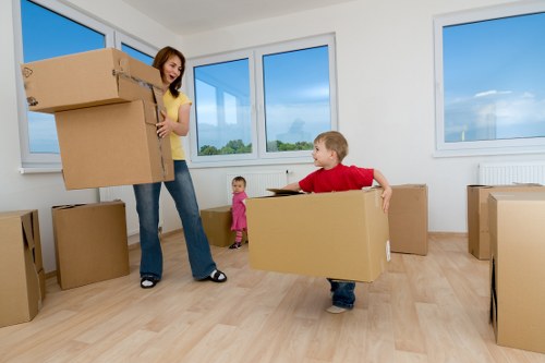 A woman in a yellow top and jeans is in a bright, spacious room holding a large cardboard box with a smile on her face. A young boy dressed in a red shirt and dark jeans is nearby, carrying a similar large cardboard box and looking up at the woman, demonstrating a collaborative move typical during house removals. In the background, a small girl in a pink outfit stands observing the activity. The room has light wooden flooring and white walls, with three large windows allowing natural daylight to fill the space. Outside the windows, a blue sky and greenery are visible, suggesting a residential setting. Several other cardboard boxes of varying sizes are placed around the room, some on the floor and one on the left side near the woman. The scene reflects a family undertaking a house move in a suburban or neighbourhood area, consistent with professional removals services such as those offered by Watson Rremovals London, focusing on efficient packing and moving within London or nearby areas like the postcode area.