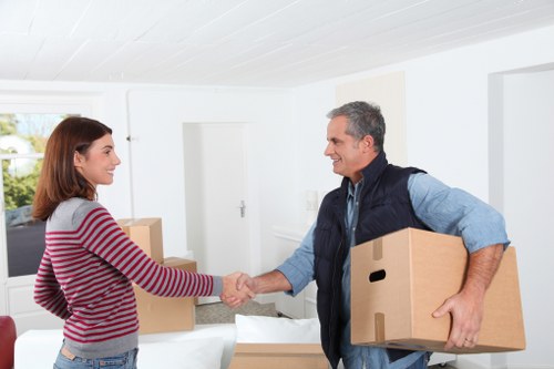 In a bright, modern interior of a residential home, a house removal specialist from Watson Removals London is seen engaging with a young woman in a casual striped sweater. The specialist, with short grey hair and wearing a blue shirt beneath a dark vest, is smiling as he extends a hand for a handshake while holding a medium-sized cardboard moving box in his other arm. The woman, with shoulder-length brown hair, is returning the handshake and has a friendly expression. Several cardboard boxes of various sizes, some sealed and others open, are visible on a table or counter behind them, indicating an ongoing move. The room features white walls, a large window allowing natural light, and a minimalist design with a plain white door in the background. The scene captures a moment of professional service and positive interaction during a house move, subtly referencing property relocation services in Londonâ€™s residential areas.