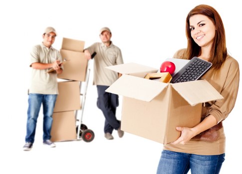 A young woman with long brown hair, dressed casually in a beige top and blue jeans, smiling while holding an open cardboard box filled with various household items, including a red apple, a black remote control, and a small yellow object. Behind her, two men are standing with additional boxes; one man is dressed in a light-colored shirt and cap, holding a stack of boxed belongings, while the other, also in a light shirt and cap, is using a hand truck to transport more boxes. The background is plain and white, suggesting an indoor or professional removals setting typical of house relocation services in Mill Hill. The scene depicts a typical moment during a house move, with the boxes neatly packed and organized, conveying a professional and efficient approach to residential removals, aligned with services offered by Watson Removals London.