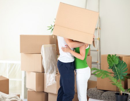 A man and woman are standing indoors surrounded by several cardboard moving boxes. The man is holding a large box in front of his head, obscuring his face, while the woman is holding the box with both hands and is close to him. The boxes vary in size and are stacked on the floor and against the wall. Some boxes are sealed with packing tape, and one appears to be wrapped in protective plastic. The room has a light-colored wall with a window partly visible on the left, allowing natural daylight to illuminate the scene. There is a grey upholstered chair with a decorative cushion on the right, partially covered by a large potted plant with broad green leaves. The scene suggests a home preparing for a house move, with the individuals engaged in a packing or moving activity, consistent with the services offered by Watson Rremovals London in the area surrounding [POSTCODE] and [TOWN].