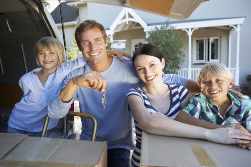 A family of four, consisting of a father, mother, and two children, are outdoors next to a house in a suburban neighbourhood. The father, with light brown hair and a smile, is holding a set of keys and standing beside a partially loaded removals van with its back door open. The mother, with dark hair and a striped shirt, is leaning on a cardboard box on a table, smiling at the camera. The children, a girl with blonde hair and a boy with blonde hair, are positioned on either side of the adults, both smiling cheerfully. The girl is wearing a light blue dress, and the boy has a checkered shirt. In the background, there is a residential house with a porch, white framing, and windows, along with greenery, suggesting a peaceful neighbourhood in London or across the UK. The scene captures the moment of a household move, with packing materials and boxes visible, reflecting a professional house removals service from Watson Removals London, situated in the postcode area.