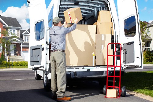 A man dressed in a light grey shirt, beige trousers, and a grey cap is shown loading cardboard boxes into the back of a white removals van parked on a residential street in Bellingham. The van's rear doors are open, revealing an interior filled with various packed cardboard boxes of different sizes, some with visible tape and labels, ready for a house move. A red hand truck or dolly is positioned upright beside the van, with a couple of small cardboard boxes resting at its base. The surroundings include a suburban area with modern detached houses featuring brick and weatherboard exteriors, manicured lawns, and a clear blue sky, suggesting good weather conditions suitable for a local house removal. The scene captures a professional and organized moving process undertaken by Watson Removals London, with attention to detail in handling household belongings for a relocation within Bellingham or nearby areas, emphasizing the company's house removal services.