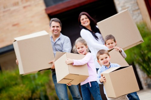 A family of four, including a man, woman, and two children, stands outdoors in front of a residential building with a brick wall and a garage door, during daytime. They are holding medium-sized cardboard boxes, which are sealed and appear to contain household items, and are smiling towards the camera. The woman has dark hair, is wearing a white long-sleeved top, and holds a box in her right hand. The man has short dark hair, is wearing a light blue shirt and jeans, and carries a box in his left hand. The two children, a girl dressed in a pink top and the boy in a striped shirt, are each holding a box, with the girl hugging her box. The background shows a grassy area and the edge of the driveway leading to the garage. This scene depicts a typical family moving day, fitting within the context of house removals services in Cowley, as offered by Watson Removals London, highlighting the process of packing and organizing household belongings for a residential move in London.