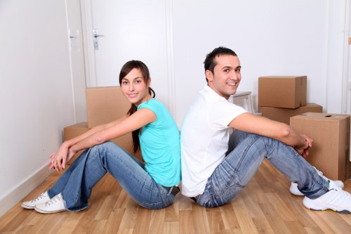 A young woman and man sit back-to-back on a light wooden floor in a bright, clean room typical of a residential property in Barnsbury, with neutral white walls and a white door in the background. The woman, dressed in a turquoise short-sleeve top and blue jeans, has long brown hair and is smiling gently, while the man, wearing a white T-shirt and grey jeans, is smiling openly. Nearby, there are several cardboard moving boxes stacked against the wall, some sealed with brown packing tape, indicating a household relocation process. The environment appears organized and tidy, with natural lighting illuminating the scene, suggestive of daytime. This setup reflects a typical stage in a house move, where belongings such as clothing and personal items are being packed in preparation for a move, managed efficiently by Watson Removals London, serving residents in the Barnsbury area, as referenced on the webpage for top-quality moving services in the postcode district associated with London N1.