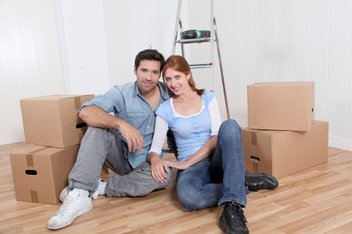 A young couple sitting on a light wooden floor inside a bright, white-walled room surrounded by packed cardboard boxes of various sizes, with some stacked on the floor and others placed on a metal step ladder in the background. The man has dark hair and is wearing a casual blue shirt, grey jeans, and white sneakers, while the woman has red hair and is dressed in a light blue shirt, dark jeans, and black boots. They are smiling and holding hands, with the woman touching her partnerâ€™s knee. The room appears to be a residential space prepared for moving, and the scene reflects the process of household relocations, compatible with services offered by Watson Removals London in the London area.