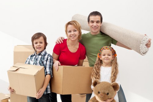A family of four, consisting of a father, mother, and two children, are standing in a bright, clean interior space filled with cardboard moving boxes. The father, wearing a green t-shirt, is holding a large rolled-up beige carpet over his shoulder, indicating a house move. The mother, dressed in a red top, has her arm around the son, who is holding a smaller cardboard box. The daughter, wearing a white top and a colorful headband, is holding a plush teddy bear. The scene suggests a positive, organized moving day, with all family members appearing happy and engaged. The background is plain white, emphasizing the household items involved in a typical house removal, including cardboard boxes of various sizes, some stacked, and a rolled carpet, aligned with the services of Watson Removals London in the Hounslow area. The setup illustrates a typical interior hallway or living room prior to relocation, highlighting the equipment used during home removals inside a residential property.