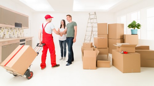 Inside a well-lit, modern home, a removal specialist from Watson Removals London, dressed in red work overalls and a red cap, is shaking hands with a male customer while a woman stands nearby, all smiling. The room features large windows allowing natural light to fill the space. To the left, a sturdy trolley loaded with cardboard boxes is positioned on light-colored wooden flooring. On the right, there are several stacks of uniformly sized cardboard boxes, some sealed with packing tape, others open with packing materials visible. A tall, aluminium step ladder leans against the wall near a small potted plant with green foliage, adding a touch of decor to the otherwise cluttered scene. The background shows a kitchen area with beige cabinetry and a sleek worktop. The setting appears to be in a residential property in Barnet, ready for a professional move. The overall environment suggests the final stages of packing or handover before a house relocation, with clear evidence of household belongings prepared for transportation, supported by Watson Removals London's experienced team in the Barnet area.