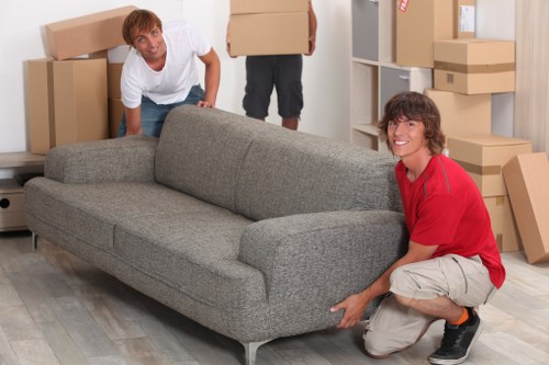 In a residential interior, two movers are shown handling a grey fabric sofa with a textured finish, positioning it carefully in a room filled with cardboard moving boxes of various sizes. One mover, wearing a white shirt, is behind the sofa, leaning forward and holding the furniture, while the other, dressed in a red shirt and beige shorts, is kneeling on the light wooden floor, supporting the front of the sofa. The room appears to be part of a house in Elephant and Castle, evident from the casual setting and the wall-mounted shelving unit partially visible on the right side, filled with more boxes and household items. The environment is well-lit, likely by natural light, with a clean and organized space dedicated to packing and moving preparations. The scene documents a professional house removal process as part of a domestic move, highlighting the careful handling of soft furnishings and household belongings by Watson Removals London, with a focus on their removals services in the local area.
