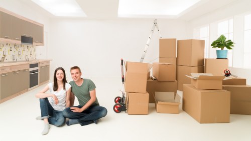 A young couple sitting cross-legged on a clean, bright kitchen floor with light-colored cabinets and a tiled backsplash. Behind them, there are several open and closed cardboard boxes of various sizes, some stacked on top of each other, indicating a recent move within a residential property. A red and black hand truck leans against the boxes, ready for use. To the right, a large window allows natural light to fill the space, illuminating a potted green plant placed near the window. In the background, a white ladder is positioned against the wall, suggesting ongoing unpacking or arrangements. The environment appears to be an interior of a house or apartment in Mayfair, with a modern and tidy setting, conveying the process of house removals with a focus on household furniture, including boxes and small items, and the preparatory stage of moving house with professional assistance from Watson Removals London.