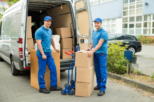 Two removalmen dressed in blue uniforms and matching caps are engaged in a moving process outside a residential building in London, with a modern multi-story structure in the background. They are loading or unloading medium-sized, light brown cardboard boxes onto or from a white moving van parked on a paved driveway. The van's open rear doors reveal more packed boxes inside. One removalman is holding a box while standing beside a hand truck or dolly, which is positioned near several stacked boxes on the ground. The other is standing on the opposite side of the boxes, smiling at the camera with a relaxed posture. In the surrounding area, there are black cars parked along the street, a small patch of green shrubbery and flowering plants near the sidewalk, and a blue mailbox. The scene captures a professional house removal operation typical of Watson Removals London, with clear focus on household items being prepared for transport, set in a residential environment during daylight hours.