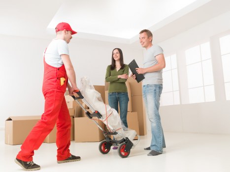 In a bright, spacious interior typical of a residential property in Archway, a professional removals team from Watson Removals London is engaged in a house removal process. The image shows a mover dressed in red work overalls and a matching cap, with a tool pouch attached to his waist, standing on the left side while handling a trolley that is supporting a large, wrapped household item. The item appears to be covered in white protective padding or blankets, indicating careful packing for transport. In the foreground, a woman with long dark hair, wearing a green long-sleeve top and blue jeans, is standing confidently, observing or discussing the move with a man on the right side, who is holding a black clipboard and dressed casually in a light grey t-shirt and blue jeans. Behind them, several cardboard boxes of different sizes are stacked, ready for loading. The natural light from large windows enhances the clean, modern atmosphere, with minimal furnishings visible, reflecting a typical move-in or move-out scenario in a residential setting in London, subtly connected to local house removal services.