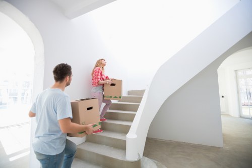 In a bright, modern interior, two individuals are engaged in a house removal process, likely part of a professional service by Watson Removals London. A woman dressed in casual pink and white checkered shirt and jeans is seen carrying a small cardboard box up a flight of curved, white-painted stairs with a textured, neutral-toned carpet. She appears to be placing the box on the staircase while holding another box, which is larger and appears to contain household items or belongings with a cardboard finish. A man, dressed in a light blue T-shirt and jeans, stands at the bottom of the stairs holding a medium-sized cardboard box and a smaller item, possibly a book or a box of miscellaneous items. The stairwell is located within a residential home on Highams Park, evident from the clean, minimalist interior design and ample natural light streaming through large, arched windows on the left, illuminating the scene. Visible in the background are white walls, a curved staircase railing, and a spacious entrance area, all contributing to a professional and organized household environment suitable for a house removals scenario.