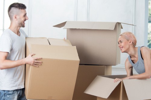 A man and a woman, both smiling, are engaged in packing or unpacking large cardboard moving boxes in a bright, spacious room with white walls and large windows. The man is standing on the left, dressed casually in a light grey t-shirt and jeans, holding a box with both hands. The woman, on the right, with platinum blonde hair styled in a bun, is leaning over a box on a low surface, wearing a sleeveless grey top. Several open cardboard boxes of varying sizes are positioned around them, some stacked, others being prepared for packing or unpacking. The scene suggests a domestic setting during a house move, with natural daylight illuminating the room, creating a professional yet casual atmosphere typical of Watson Removals Londonâ€™s house removals service in the London area, possibly near South London or the surrounding postcode district. No additional furniture or equipment is visible in the scene, focusing solely on the boxes and the individuals involved in the moving process, reflecting a moment of preparation for a residential move with attention to detail and care.