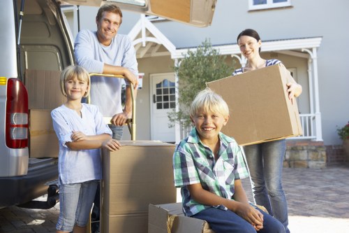 A family of four, including a man, woman, and two children with blonde hair, are outside their residential house during daytime engaging in household moving activities. The man, wearing a light blue shirt, is standing in the back of an open removals van and holding a yellow strap while smiling at the camera. The woman, dressed in a striped top, is holding a large cardboard box and standing next to the van. The two children, a girl and a boy with light-colored hair, are positioned in front: the girl, in a light blouse and jeans, is standing next to a stack of cardboard boxes on the driveway, and the boy, in a green and blue checkered shirt, is sitting on a box at ground level, smiling at the camera. The driveway is paved, and in the background, there is a modern two-storey house with white siding, a grey roof, large windows, and some greenery, indicating a suburban area that could be in London or nearby. The scene is well-lit with natural daylight, capturing a typical house removal moment, illustrating the process of packing and loading belongings with professional service from Watson Rremovals London, whose branding connects naturally with residential moving services in the area.