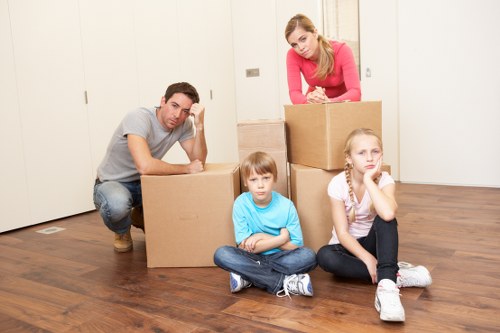 A family of four, consisting of a man, a woman, a young boy, and a young girl, is inside a residential property preparing for a move. The man, dressed in a grey T-shirt and jeans, leans on a large cardboard box placed on a wooden floor, appearing contemplative. The woman, wearing a pink top, stands behind a stack of three cardboard boxes, looking down at the camera with a neutral expression. The young boy, sitting cross-legged in front of the boxes, is wearing a light blue T-shirt and denim jeans, with a serious look on his face. The young girl, sitting on the floor next to the boy, has a thoughtful expression, resting her chin on her hand, and is dressed in a white shirt and black trousers. The background features white panelled walls and a closed door, indicating a tidy interior space typical for a household in Stanmore. The scene captures a moment of packing or organizing, characteristic of house removals services offered by Watson Removals London, with focus on household furniture, moving boxes, and personal belongings in preparation for a move within London or surrounding areas.