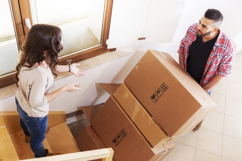 A professional removals team from Watson Removals London assists a woman inside a residential property in Stroud Green, London, with moving large cardboard boxes. The scene takes place near the entrance, showing the woman standing on a wooden staircase, gesturing towards the boxes, which are stacked and held by a man wearing a red plaid shirt. The boxes are sealed with packaging tape and feature handling symbols, indicating they contain household items. The interior environment includes a white wall, a window with a wooden frame, and a tiled floor, suggesting a welcoming and well-maintained home in the area. The lighting appears natural, likely coming from the window, illuminating the scene clearly. This image captures the careful handling and organized process involved in house removals, illustrating the services offered by Watson Removals London in the Stroud Green area.