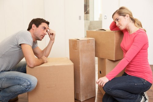 A man and a woman are indoors in a residential property, surrounded by several stacked cardboard moving boxes in various sizes, some of which are open. The man, dressed in a light grey t-shirt and blue jeans, is sitting on the floor with his left arm resting on a box while touching his forehead with his right hand, appearing contemplative. The woman, wearing a bright pink long-sleeved top and dark jeans, is seated on the floor leaning against a large cardboard box with her head resting on her arm, looking thoughtfully at the man. The background shows a modern, well-lit interior with neutral-colored walls and a staircase leading upward, suggesting a home environment typical of a house in Stroud Green or nearby London districts. The scene captures a moment during a house move, with furniture and household items not visibly present but implied by the boxes and setting, reflecting professional removals services by Watson Removals London in the area.