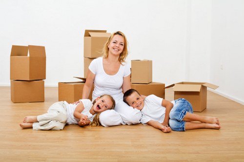 A smiling woman with shoulder-length blonde hair, dressed in a white t-shirt and light-colored pants, sits on a wooden floor in a bright, minimally furnished room with plain white walls. Surrounding her are several cardboard moving boxes, some stacked behind her and others open or closed, indicating a house move or packing process. On her left, a young girl with long blonde hair, wearing a white top and beige pants, lies on her stomach, resting her head on her hand and smiling cheerfully. To her right, a boy with short dark hair, dressed in a white t-shirt and blue jeans, lies on his side, propped up on one elbow, also smiling. The scene has a warm, natural lighting, emphasizing a cheerful and organized environment typical of a family preparing for or during a home relocation, with a focus on house removals services in London. The background shows a clean, plain white wall with no additional furniture or decor, highlighting the moving boxes and the relaxed, happy family atmosphere.