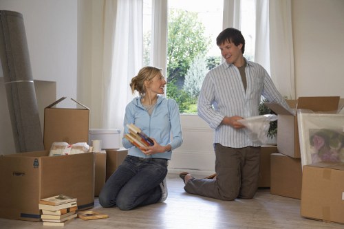 A woman and a man are inside a well-lit, spacious room with large windows showing green trees outside. The woman, with blonde hair tied back, is kneeling on the wooden floor, holding a stack of books and smiling at the man. The man, with short dark hair, is standing next to several open cardboard boxes filled with various household items, including a pink object and plastic wrapping. He is wearing a light striped shirt and dark trousers. The room contains additional packed boxes stacked against the wall, some with visible packing materials, and a few books and small objects scattered on the floor. Natural daylight illuminates the space, highlighting the textures of the wooden flooring, cardboard boxes, and fabric of their clothing, reflecting a typical home setting during a house move in Tokyngton. Watson Removals London appears to be assisting in a professional and organized manner, preparing for a relocation or house clearance at a residential property.