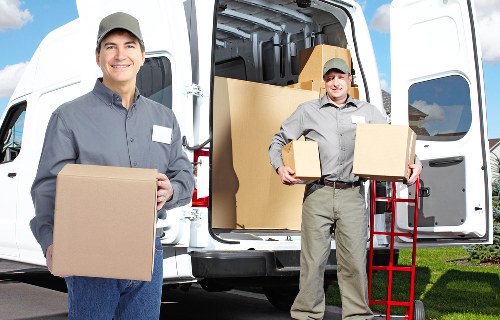 Two professional house removals staff members are positioned outdoors near a white removals van, which has its rear doors open, revealing several packed cardboard boxes and large moving cartons. The staff are dressed in grey work uniforms with caps, and they are actively engaged in lifting and carrying boxes; one staff member is holding a medium-sized box in front of him, smiling at the camera, while the other is carrying a larger box and standing beside a red hand trolley loaded with additional boxes. The scene takes place on a paved driveway adjacent to a residential property, with a well-maintained lawn and some greenery visible in the background. The interior of the van shows a wall of stacked boxes and packing materials, indicative of a professional moving operation in Grove Park. The environment appears bright and clear, suggesting daytime, and the focus is on the safe handling and transportation of household belongings by Watson Removals London, supporting efficient and reliable household relocation services in the local area.