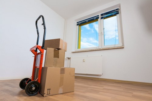 A room with plain white walls, featuring a large double-glazed window with blue and yellow window frames, allowing natural daylight and showing an outdoor view of a partly cloudy sky. In the foreground, there are three cardboard boxes stacked on top of each other, with the largest at the bottom, all sealed with packing tape and positioned near a white radiator mounted on the wall underneath the window. To the left of the boxes, a red hand truck with black wheels and handle is leaning against the boxes, ready for moving or transport. The room has a light wooden floor, and no furniture or other objects are visible, indicating the space is either freshly cleared or prepared for a move, consistent with professional house removal services in Pimlico as provided by Watson Removals London. The scene captures a quiet, organized interior typical of an ongoing move within a residential property.