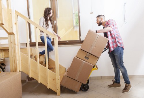 A man and a woman are engaged in a house removal process inside a residential property. The man, wearing a red and white checkered shirt and jeans, is pushing a small, black, wheeled trolley loaded with three large cardboard packing boxes, one stacked on top of another. He is in the act of guiding the trolley towards the staircase, which has light wooden steps and matching handrails. The woman, sitting on the wooden staircase and dressed in a white top and blue jeans, is helping to steady or receive the boxes. The interior features a beige tiled floor and white walls, with a large window letting in natural light to illuminate the scene. In the background, there is a partially visible door or glass panel leading outside or to another room, indicating this is a lively, ongoing moving or packing operation typical of services offered by Watson Removals London. The environment suggests a tidy, well-maintained home in Coombe, with the focus on careful handling and organization of household belongings during a professional house move.