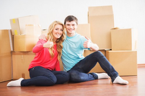 A young couple with light skin and casual clothing, including a woman in a red sweater and a man in a light blue t-shirt, are sitting on a wooden floor inside a residential property in Maze Hill, surrounded by multiple cardboard boxes of various sizes, some sealed with packing tape. The boxes are arranged behind them, indicating they are in the process of packing or preparing for a house move. The room has a light, neutral-colored wall, and the lighting appears natural and bright. Both individuals are smiling warmly, with the woman giving a thumbs-up gesture and the man holding a small box, suggesting a positive mood related to house removals or moving activities. Nearby, a white door is visible, and the scene exudes a sense of organized activity associated with home relocation by Watson Removals London, serving clients in the Maze Hill area. The overall impression is one of a cheerful, carefully packed home interior ready for a move, reflecting professional moving services within a residential setting.