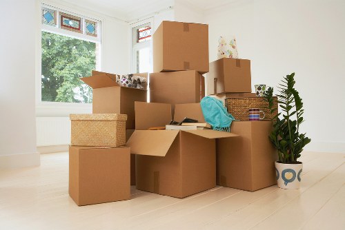 A spacious, well-lit room with white-painted walls and light wooden flooring, filled with multiple brown cardboard moving boxes of various sizes, some open revealing contents such as clothing or small household items, while others are sealed. The boxes are stacked on top of each other and arranged across the floor, indicating packing in progress within a residential property near London. A woven basket and a blue garment or cloth are placed on one of the boxes, and a potted plant with large green leaves sits to the right, providing a touch of decor. In the background, two large windows with white frames allow natural daylight to illuminate the space, with a view of leafy trees outside. The scene depicts an interior setting associated with house removals, highlighting the process of packing and preparing belongings for a move, typical of a service like Watson Removals London operating in the postcode area around the town or district implied by the page title.