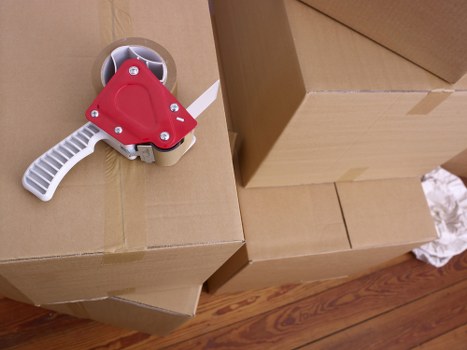 A close-up view of a cardboard box stacked in a living space, with a red and white tape dispenser placed on top of it. The box appears to be part of packing or moving materials, featuring brown corrugated cardboard with visible flaps and tape. In the background, there are more cardboard boxes, slightly out of focus, indicating an ongoing packing or moving process. The setting is within a residential area, evidenced by the wooden flooring beneath the boxes. The scene captures the preparation phase of a house relocation, typical for professional removals services such as Watson Removals London operating in Cranham, aimed at assisting with household moves within the local area. The lighting is natural and even, highlighting the texture of the cardboard and the clean, organized environment typical of a house being prepared for moving appropriate for a professional service webpage focusing on removals in the Cranham area.