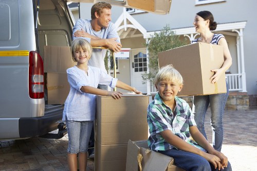 A family of four, including two children and two adults, are shown in a residential driveway preparing for a house move in Chiswick. The adults are standing near an open grey removal van, which is parked close to a white house with a pitched roof and a front garden. The woman, dressed in a striped top, holds a medium-sized cardboard box with tape along the edges, while the man, wearing a casual blue shirt, leans on the side of the van, watching the children. The children, a girl in a white top and denim shorts and a boy in a plaid shirt, are handling smaller cardboard boxes, stacking them on the driveway, which is paved with bricks. The boxes are sealed with packing tape and vary in size, some ready for transportation. The scene is well-lit, suggesting a clear day, with the family appearing cheerful and organized, preparing belongings for a local move within the London area. Watson Removals London specializes in this type of residential house removal, ensuring a smooth transition in Chiswick and broader London neighborhoods.