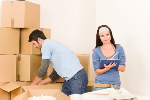 A man and a woman are involved in packing household items in a spacious, well-lit room during a house moving process. The man, dressed in a light blue t-shirt and dark jeans, is leaning forward and carefully packing a cardboard box, surrounded by several other boxes of various sizes, some sealed with tape and others open, revealing packing material and household belongings. In the background, tall stacks of filled boxes are visible against a plain white wall, indicating an organized packing effort within a residential space. The woman, wearing a light grey headband, blue long-sleeve top, and casual trousers, stands slightly to the side, holding a clipboard and pen, possibly overseeing or managing the packing process. The environment suggests a focus on house removals or relocations, with the scene captured in natural daylight, reflecting a typical domestic setting in Arkley or nearby areas, demonstrating the professional approach of Watson Removals London in handling house moves efficiently and systematically.