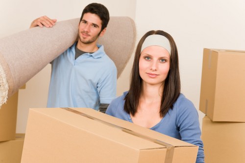 A young man with short dark hair and a light blue shirt is carrying a large, beige upholstered armchair over his shoulder inside a residential property in Motspur Park, suggesting a house removal or relocation. Behind him, a young woman with shoulder-length dark hair, wearing a grey headband and a blue top, is holding a medium-sized cardboard box, ready for transport. The background includes additional cardboard boxes, some stacked, indicating packing activities typical of a house move. The room has neutral-colored walls and appears to be a living area, with a focus on the process of loading household items. The natural lighting highlights the textures of the cardboard, fabric, and upholstery, emphasising the efficiency and organization of a professional removals service by Watson Removals London. The scene depicts the careful handling of furniture and belongings, relevant to residents in the London area preparing for relocation, and aligns with the services offered in Motspur Park postcode area.
