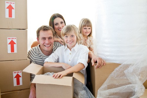 A family of four, including a man, woman, and two children, are inside a well-lit room during a house move. The children, a boy with light blond hair in a white shirt and a girl with shoulder-length blonde hair in a floral dress, are sitting in and around open cardboard boxes filled with packing material. The man, with short blond hair and a striped shirt, is leaning into a box, smiling at the camera, while the woman with long dark hair, dressed in a white top, stands behind them, also smiling. In the background, there are stacked cardboard boxes labeled with red arrows indicating the correct orientation, some partially opened, and some wrapped in protective plastic or bubble wrap. The room has bright, natural lighting, with a plain white wall and a partially visible window or curtain on the right side. The scene depicts a typical house relocation process, highlighting packing activities and family involvement, consistent with house removals services in West Brompton.
