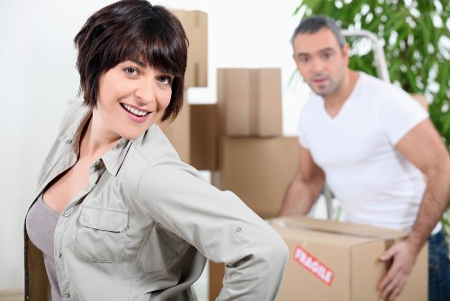 A woman with short dark hair, wearing a light grey jacket, is smiling at the camera in the foreground of an indoor residential space, with natural light illuminating her face. Behind her, a man with short dark hair and wearing a white t-shirt is slightly blurred, as he holds a cardboard box with a red and white label, beside several stacked moving boxes. The background includes a white ladder and green leafy plants, suggesting an interior room during a house relocation, likely on a residential street in Archway. The scene depicts a professional house removals process, with attention to the packing, and careful handling of household items, consistent with the services offered by Watson Removals London. The environment appears organized, with neutral colours on the walls and a tidy setting, emphasizing the careful planning involved in stress-free moving. The overall atmosphere is professional, calm, and focused on household logistics, supporting local relocation services in London.